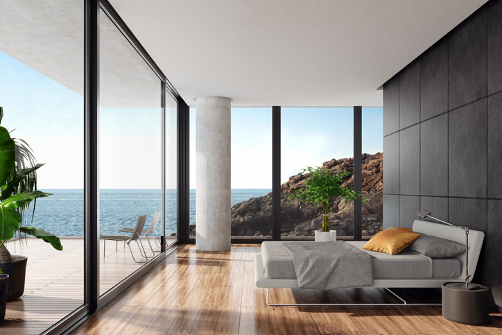 Luxurious minimalist bedroom in a seaside villa with big panoramic windows
and ocean view. White textile king size bed with white metal construction in front of 
black wall with natural stone large tiles. Round modern gray nightstand with a
modern metal lamp next to the bed. Wooden varnished floor. Big terrace with an
ocean view and glass fence with green plants. Summer daylight scene.
Background is my own photo.