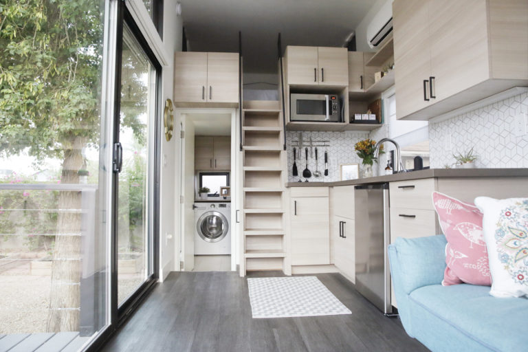 The interior of a tiny house with large glass windows, showcase the kitchen, part of the living room and loft bedroom.